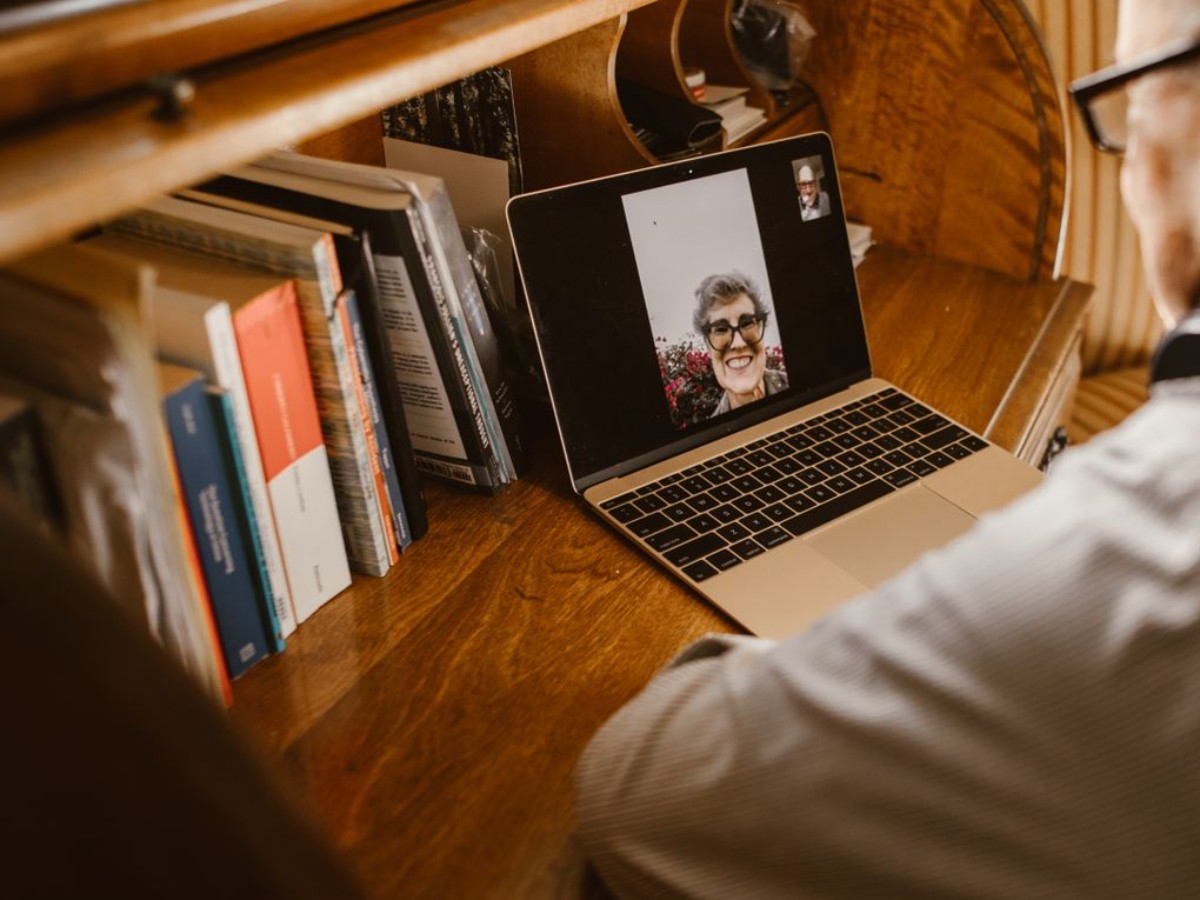 A laptop open on a wooden desk beside a bookshelf, a video call in progress on screen